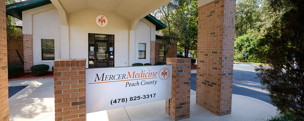Behind a Mercer Medicine peach county sign supported by two red brick columns, A door into a white building can be seen with the Mercer Medcine logo above.